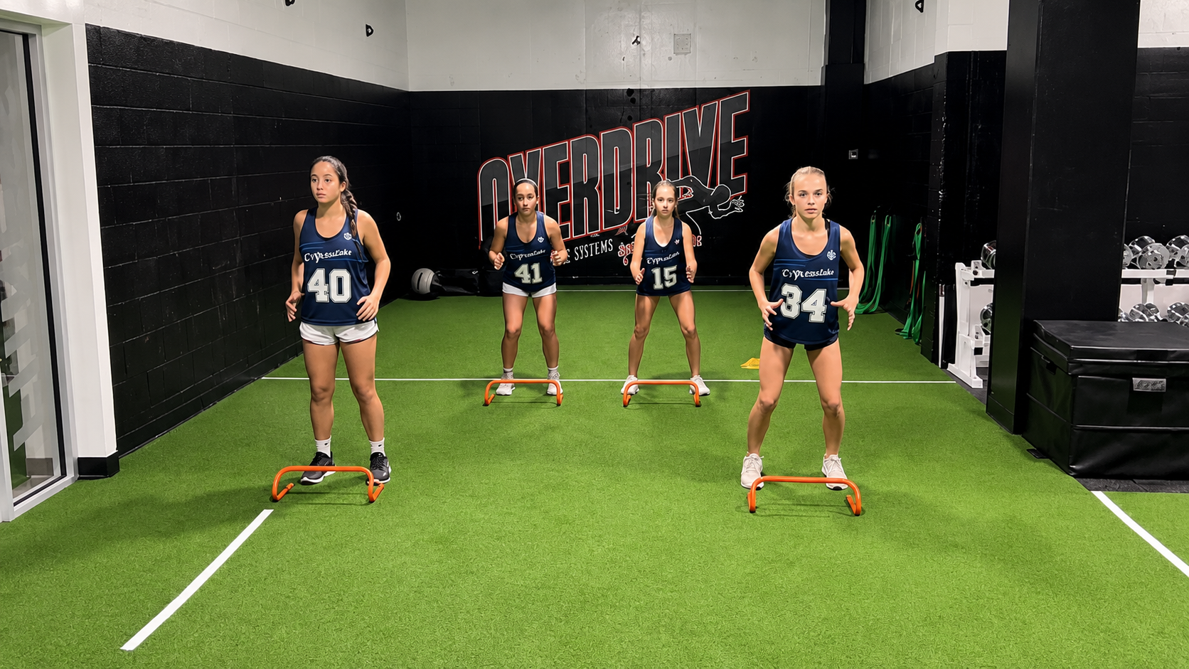 Group of young female athletes performing hurdle drills in front of the Overdrive wall logo